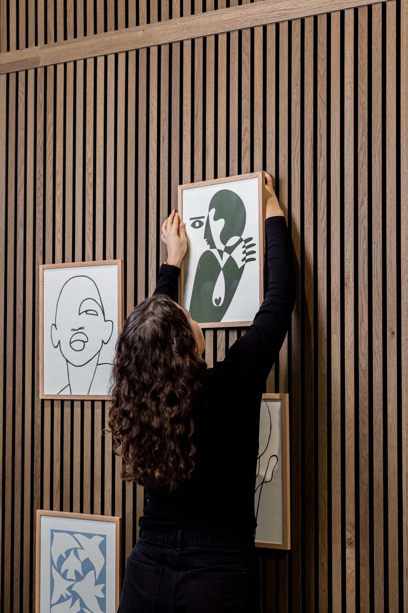 Eine Person mit lockigem Haar hängt ein WoodUpp® CREATE DUPPs Zubehör an ein akustisches Lamellenwandpaneel aus Holz, umgeben von minimalistischer Kunst und modernem Dekor.